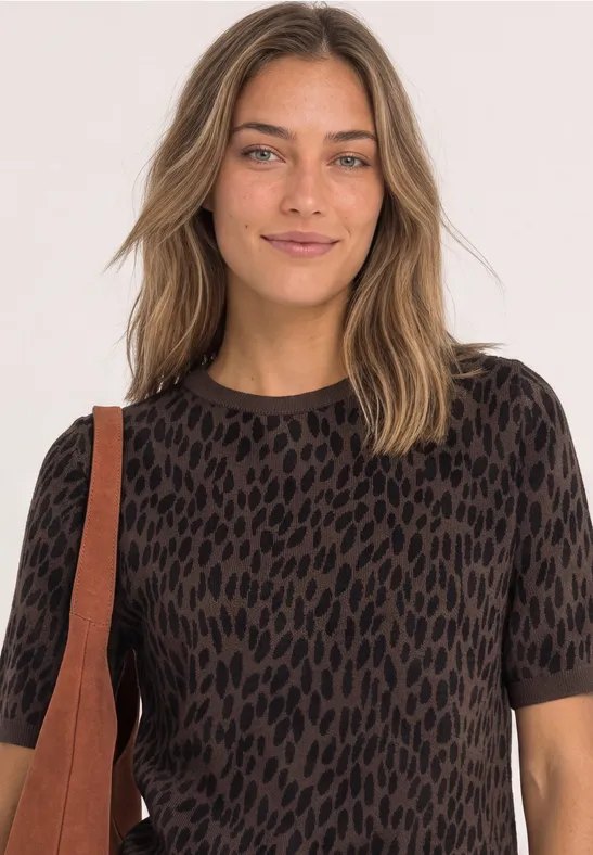 Eine Frau mit langen Haaren trägt ein kurzärmeliges, braun-schwarz gemustertes Shirt und eine Schultertasche.