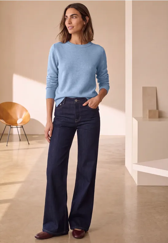 Vrouw in minimalistische ruimte draagt lichtblauwe trui, donkere wijd uitlopende jeans en bruine schoenen.
