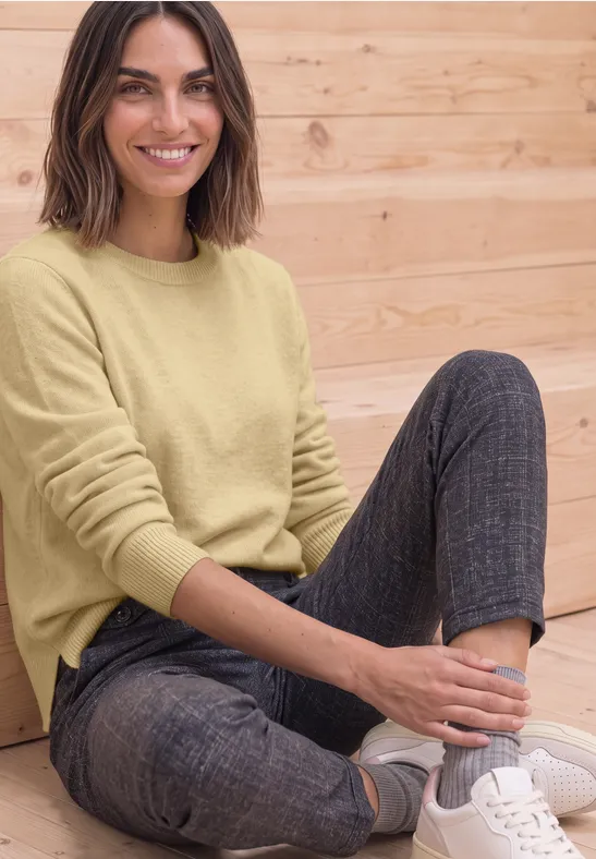 Eine Frau sitzt lächelnd vor einer Holzwand, trägt gelben Pullover, graue Hose und weiße Sneaker.