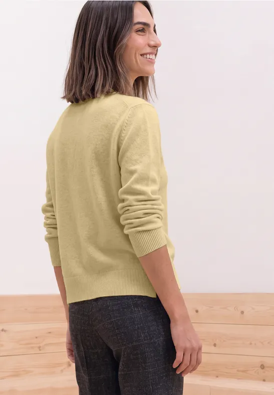 Frau von hinten in hellem Pulli und dunkler Hose, vor hellem Hintergrund.