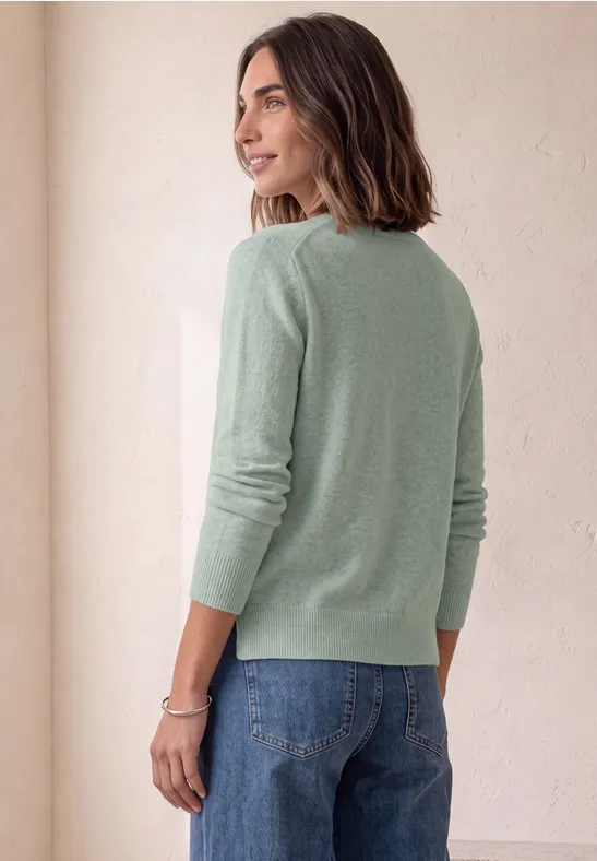 Eine Frau in grünem Pullover und Jeans steht seitlich, Blick über die Schulter, vor hellem Hintergrund.