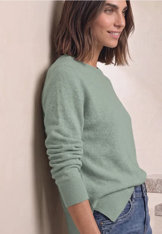 Frau lehnt an Wand, trägt grünen Pullover und blaue Jeans, lächelnd und entspannt.