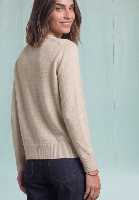 Vrouw in beige trui en spijkerbroek kijkt over haar schouder en glimlacht licht.