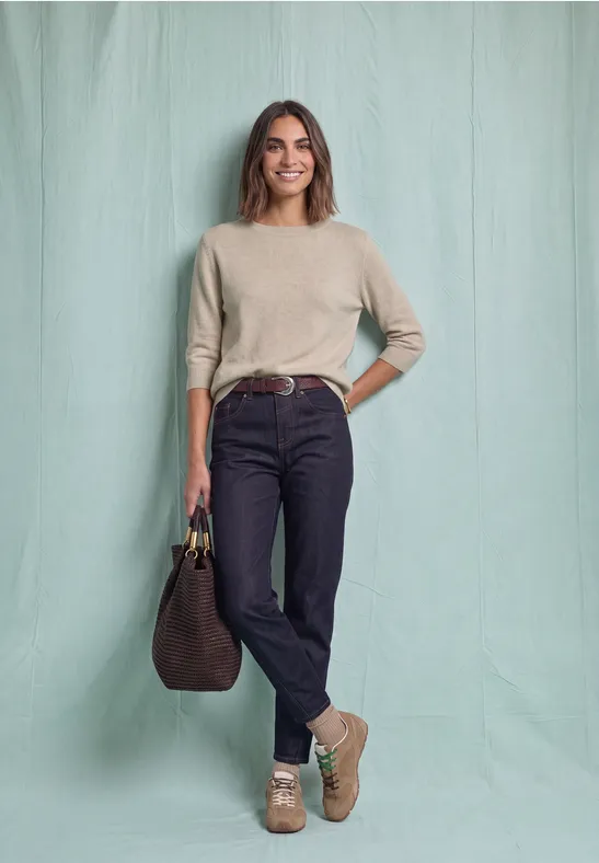 Vrouw met beige trui, zwarte jeans, lichte enkellaarsjes en grote bruine tas voor een lichtgroene achtergrond