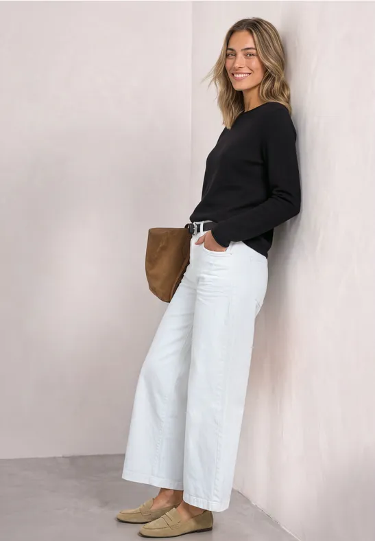 Eine junge Frau lehnt lächelnd an einer hellen Wand, trägt schwarzen Pullover und weiße Jeans, hält eine braune Tasche.