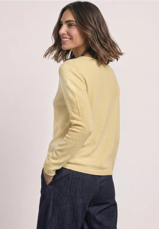 Eine Frau in gelbem Pullover und dunkler Hose blickt lächelnd seitwärts.