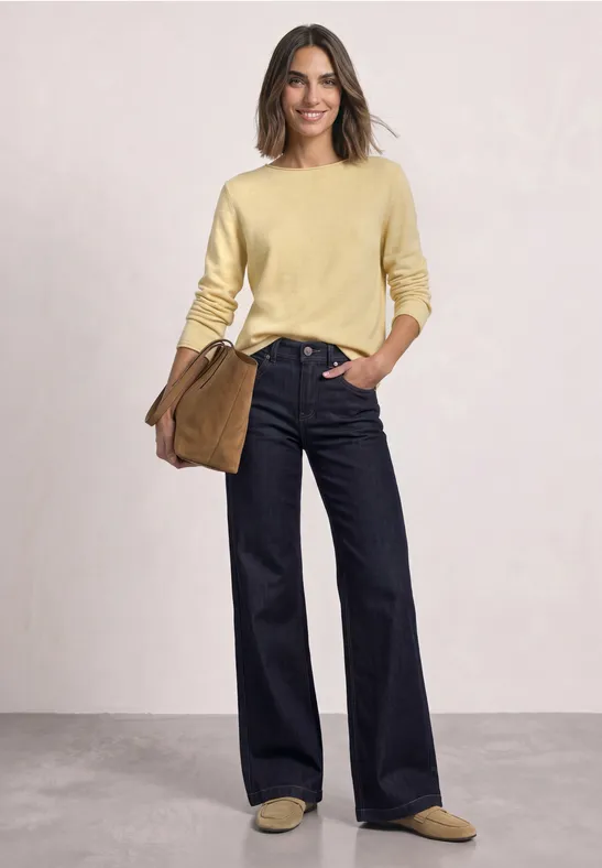 Frau in gelbem Pullover, weiter dunkelblauer Jeans, beigen Schuhen und brauner Tasche.