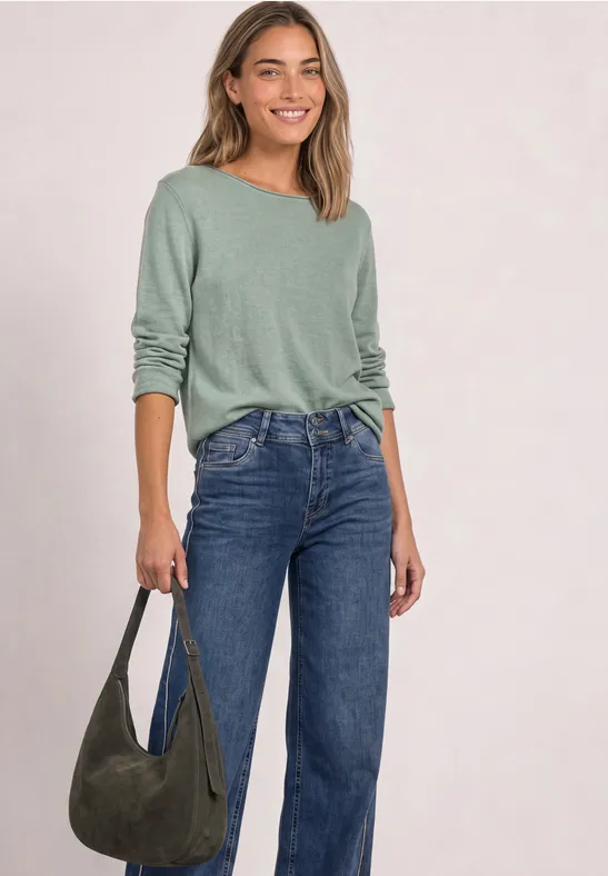 Eine Frau mit graugrünem Langarmshirt, blauen Jeans und grüner Schultertasche, die lächelnd posiert.