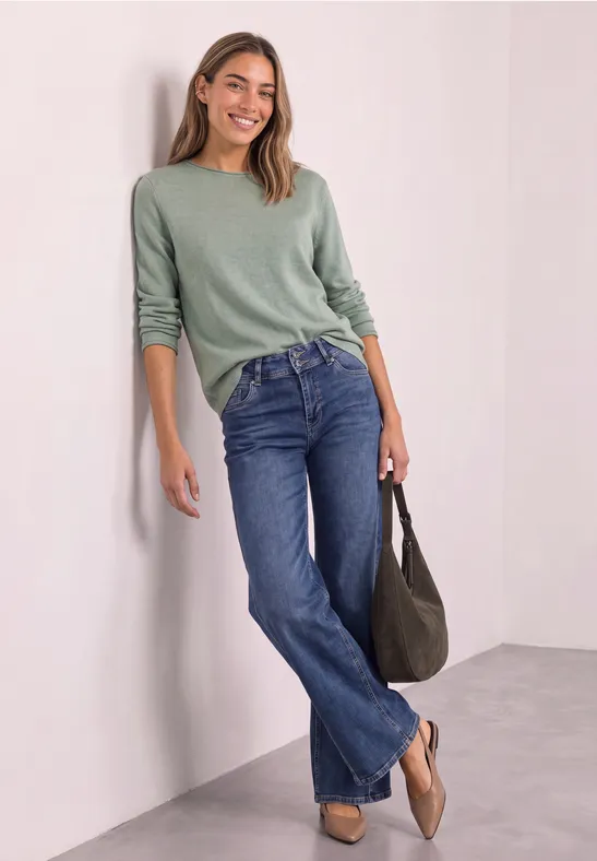 Junge Frau lehnt lächelnd an weißer Wand, trägt grünen Pullover, blaue Jeans und hält eine Tasche.