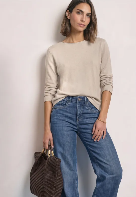 Frau in beigem Pullover und blauer Jeans mit brauner Geflecht-Tasche.