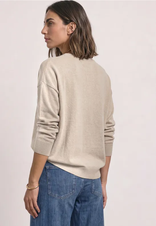 Frau von hinten in beigem Pullover und blauen Jeans vor hellem Hintergrund.