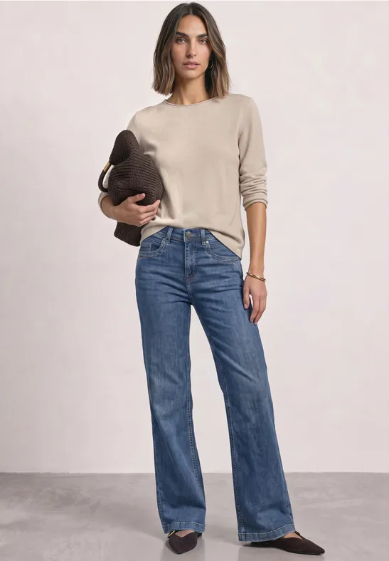 Frau in beigem Pullover und blauen Jeans, hält eine Tasche in der Hand.