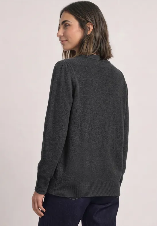 Frau im Profil mit grauem Pullover und blauer Jeans vor hellem Hintergrund.