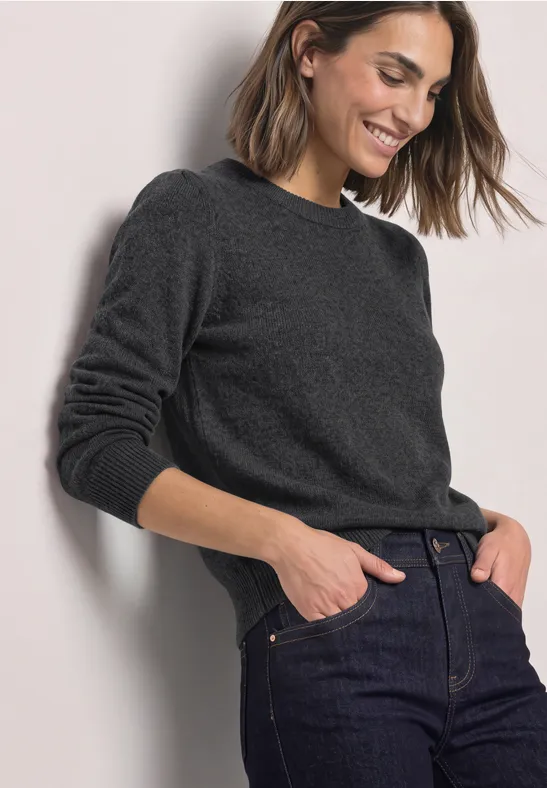 Frau mit dunkelgrauem Pullover und Jeans lehnt lächelnd an Wand.