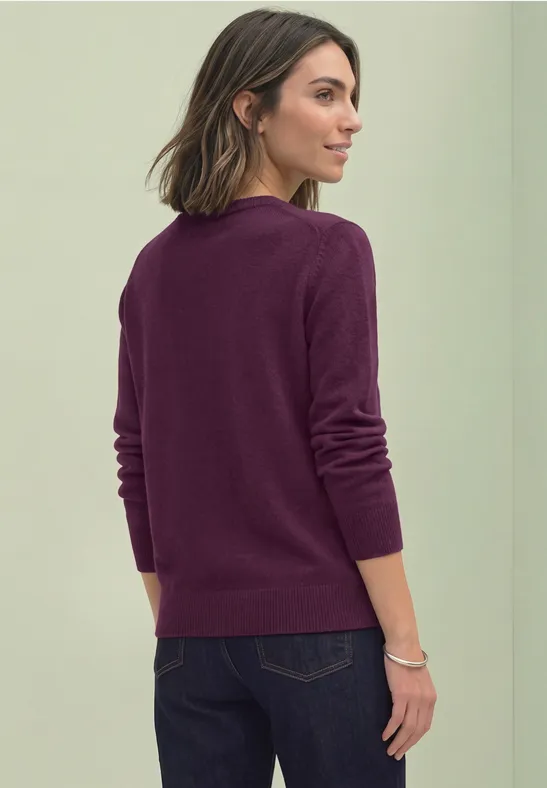 Frau in pflaumenfarbenem Pullover und dunkler Jeans, Rückenansicht vor neutraler Wand.
