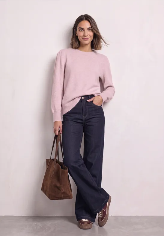 Frau in rosa Pullover, weiten Jeans und braunen Loafers trägt eine braune Tasche, posiert vor neutralem Hintergrund.