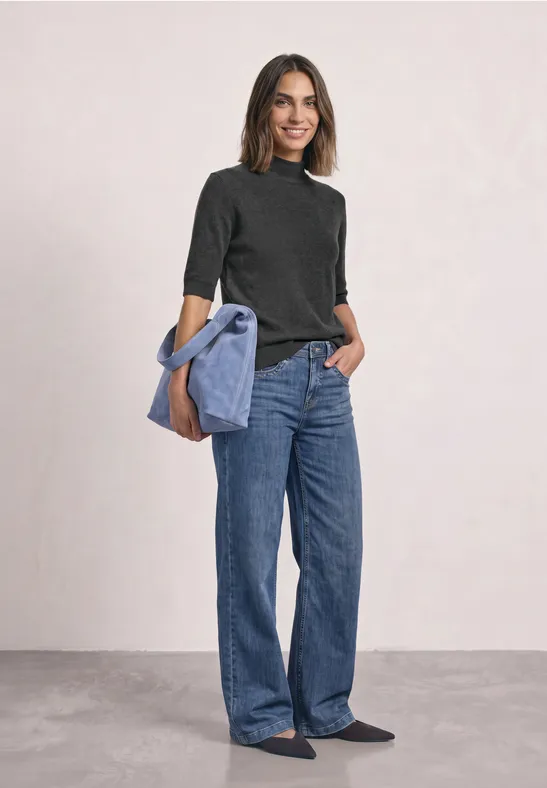 Frau in weiten Jeans und schwarzem Pulli, hellblaue Jacke lässig über der Schulter, lächelnd.