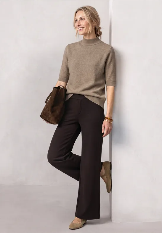 Femme en pull beige, pantalon noir et bottines vertes, souriante, appuyée contre un mur blanc.