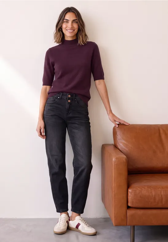 Femme appuyée sur un canapé en cuir marron, pull violet, jean noir, baskets blanches, sourire.