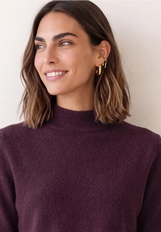 Femme aux cheveux châtains, boucles d’oreilles dorées, souriante avec un pull violet à col roulé.