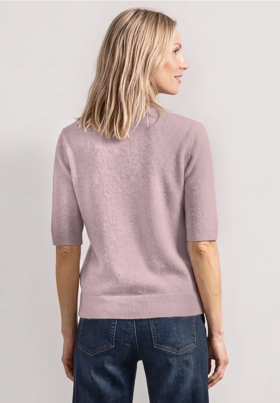Frau mit schulterlangem blondem Haar trägt rosafarbenen Pullover und Blue Jeans, von hinten fotografiert.