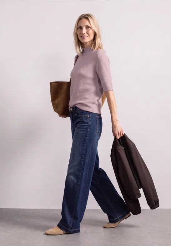 Frau in hellem Pullover und weitem Jeans, mit Tasche über der Schulter und Jacke am Arm.