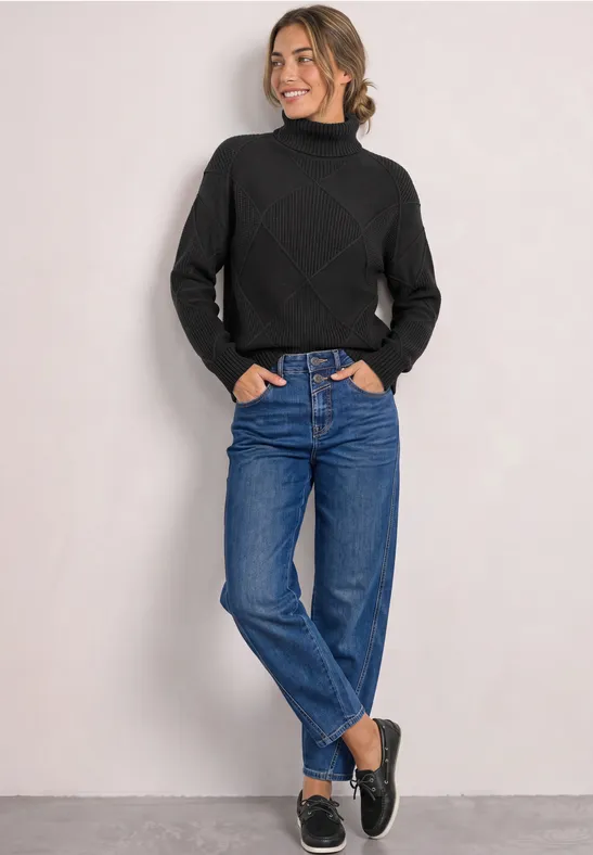 Junge Frau in schwarzem Rollkragenpullover und blauen Jeans vor neutralem Hintergrund.