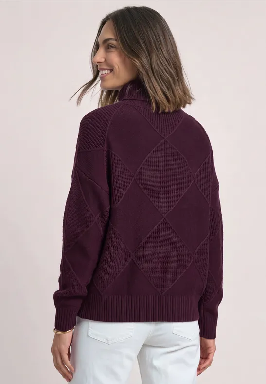 Frau im lila Strickpullover mit Stehkragen und Rautenmuster, weiße Hose, lächelt seitlich.
