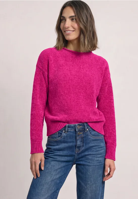 Frau trägt pinken Pullover und blaue Jeans, steht lächelnd vor hellem Hintergrund.