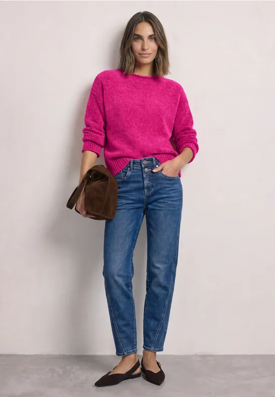 Frau in pinkem Pullover, blauen Jeans und schwarzen Ballerinas, mit brauner Clutch vor hellem Hintergrund.