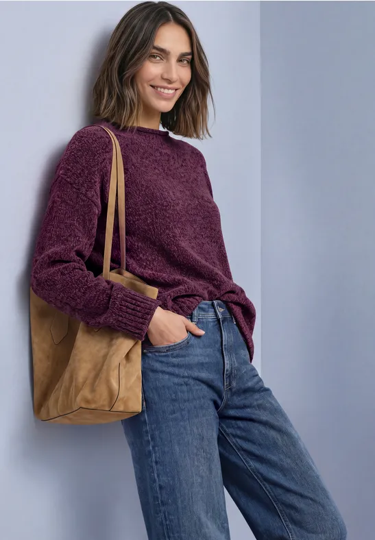Frau lehnt an Wand, trägt violetten Pullover, blaue Jeans und eine braune Umhängetasche, lächelt.