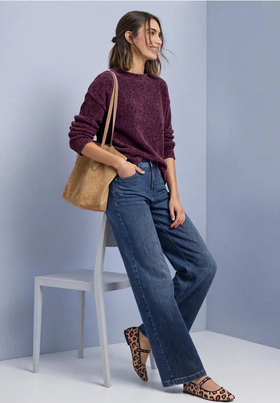 Frau in lila Pullover, weiten Jeans und Leo-Slippern mit brauner Tasche vor hellblauem Hintergrund.