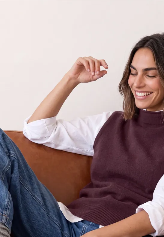 Een lachende vrouw ontspant op een bank, draagt een gilet en jeans.
