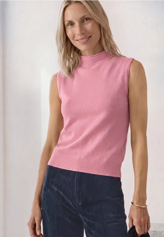 Een glimlachende vrouw draagt een roze mouwloze top en een donkere broek.