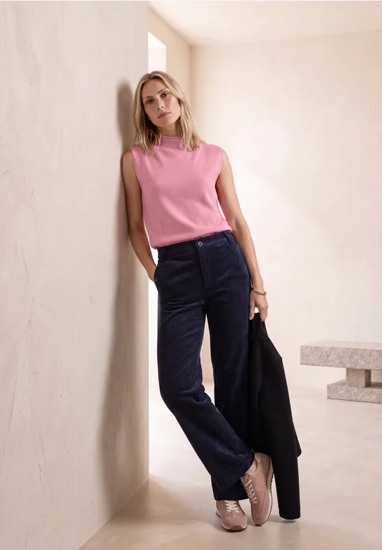 Blonde vrouw leunt tegen een beige muur, roze top, donkerblauwe corduroy broek, zwarte blazer in de hand.