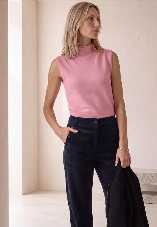 Vrouw in roze top en zwarte broek, houdt zwarte blazer, kijkt opzij.