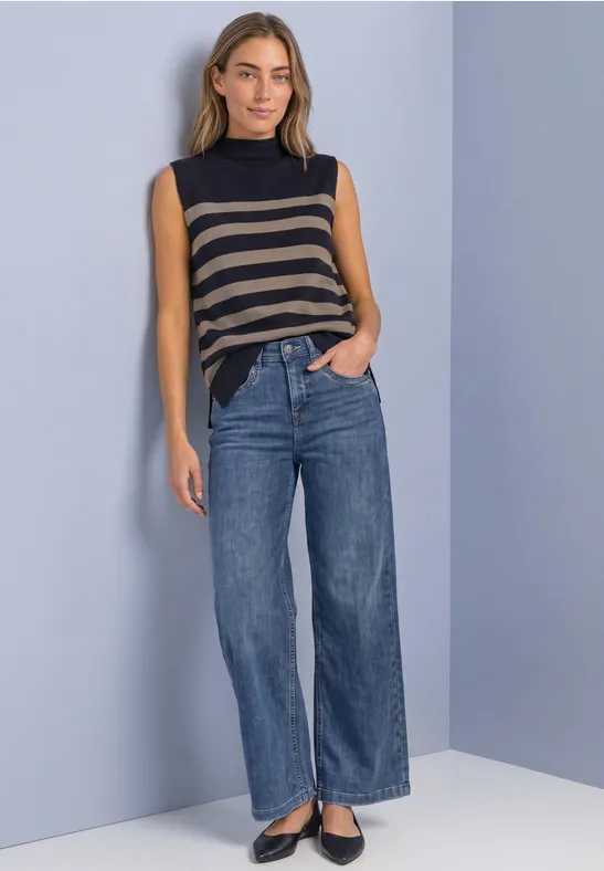 Frau im ärmellosen gestreiften Top, weiten Jeans und schwarzen Ballerinas vor schlichtem Hintergrund.
