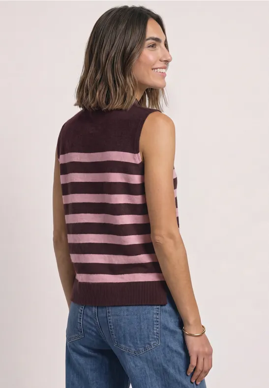 Dame im Halbprofil trägt ärmelloses, bordeauxfarbenes Top mit rosa Streifen und blaue Jeans.
