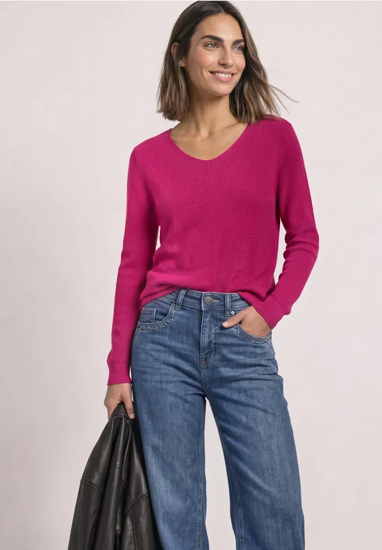 Frau in pinkem Pullover und blauen Jeans hält schwarze Tasche und lächelt.