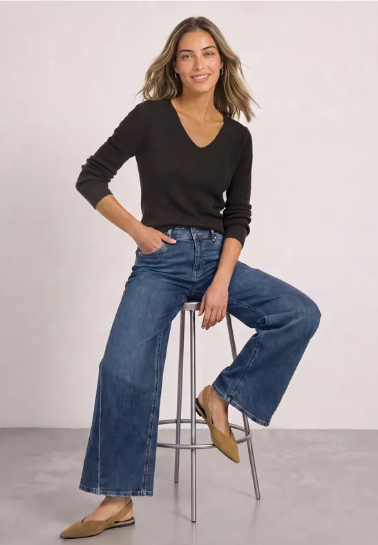 Eine Frau sitzt lächelnd auf einem Hocker, trägt schwarzen V-Pullover, weite Jeans und beige flache Schuhe.