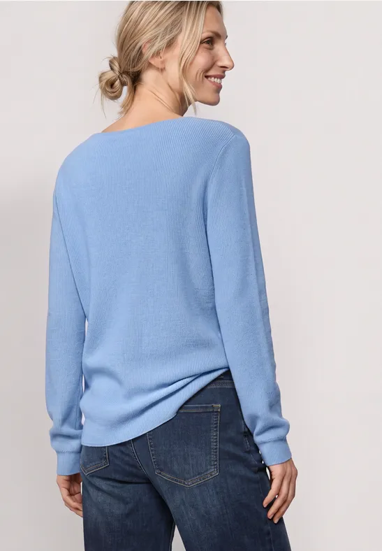Frau steht mit dem Rücken zur Kamera, trägt hellblauen Pullover und Jeans, lächelt seitlich.