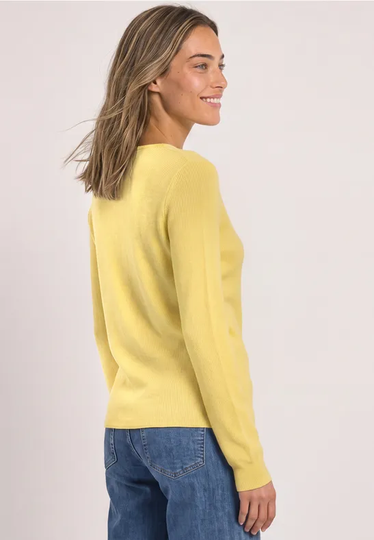 Eine Frau in gelbem Pullover und Jeans, seitlich gewandt, lächelt.