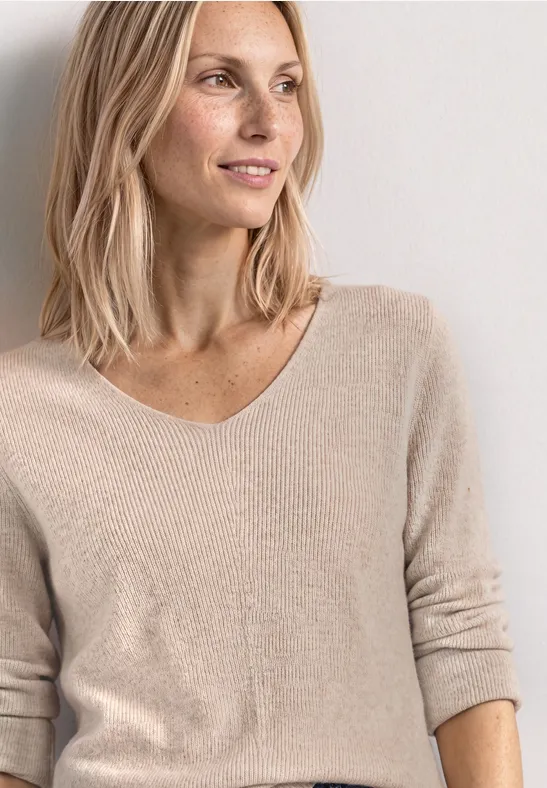 Femme blonde élancée, portant un pull beige à col V, regardant légèrement sur le côté avec un sourire.
