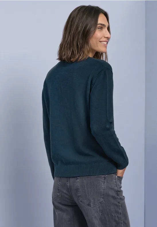 Frau von hinten, trägt einen dunkelblauen Pullover und graue Jeans vor hellem Hintergrund.