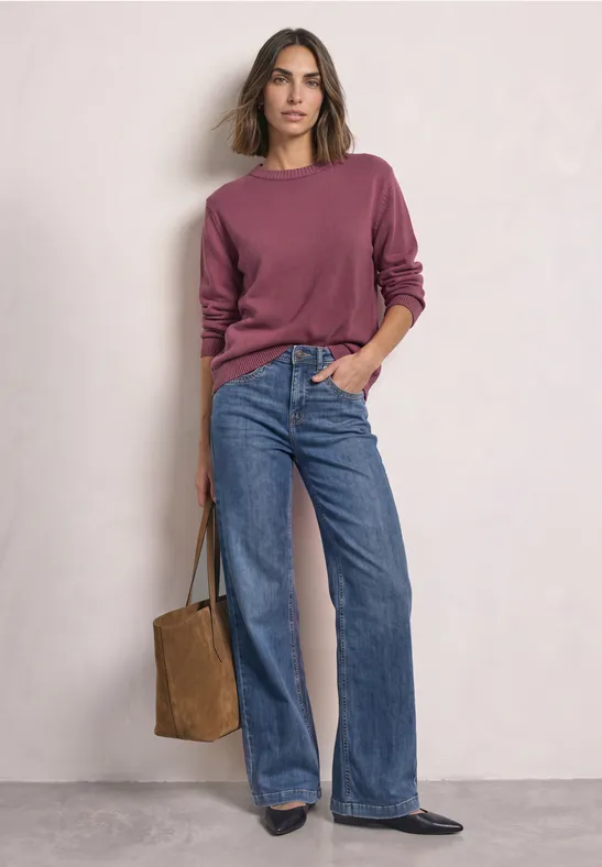 Frau steht vor weißer Wand, trägt bordeauxfarbenen Pullover, weite Jeans und hält eine braune Tasche.