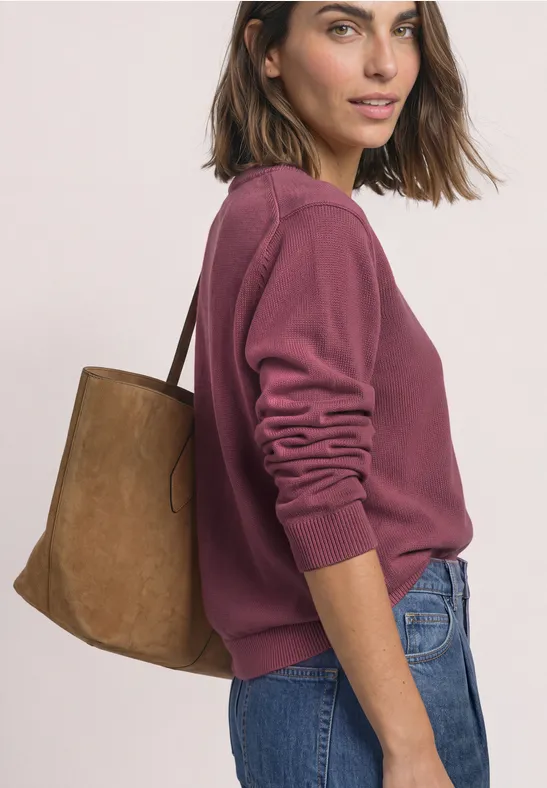 Frau in violettem Pullover und Jeans trägt eine braune Schultertasche, seitliche Ansicht.