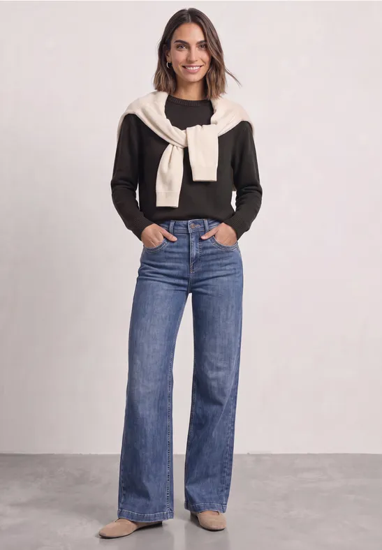Frau trägt schwarzen Pullover, beigen Pullover über den Schultern und weite blaue Jeans.