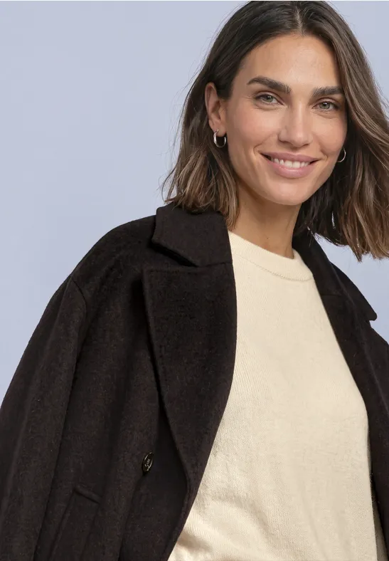 Eine lächelnde Frau mit schulterlangen braunen Haaren trägt schwarzen Mantel und hellen Pullover.
