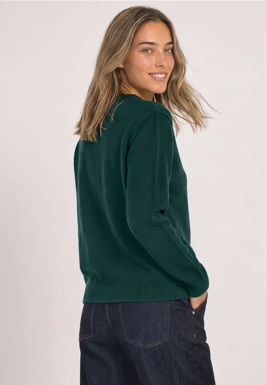 Frau in grünem Pullover und Jeans, seitlich stehend, lächelt in die Kamera.