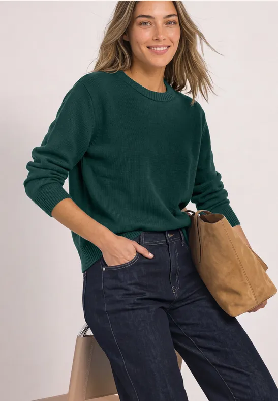 Eine junge Frau in dunkelgrünem Pullover, Jeans und beiger Tasche, die lächelnd in die Kamera schaut.